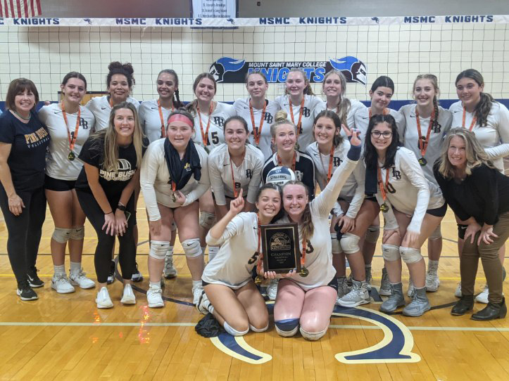 Eighteen high school girls i blue and white volleyball uniforms, and two adult women on either side. All have medals around their necks and some are holding up #1. The two in the front are holding a large plaque.