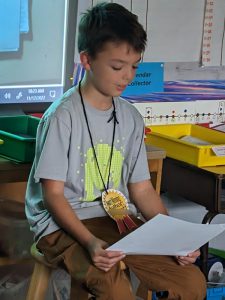A fourth-grade boy, wearing a gray shirt with a yellow design on it sits on a chair and reads from his book he wrote.