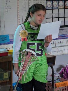 A fourth-grade girl, wearing a green lacrosse uniform with the number 55 and holding a lacrosse stick, stands and reads from the book she wrote.