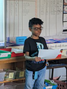 A fourth-grade boy, wearing a black shirt and glasses, stands and reads from his book he wrote.