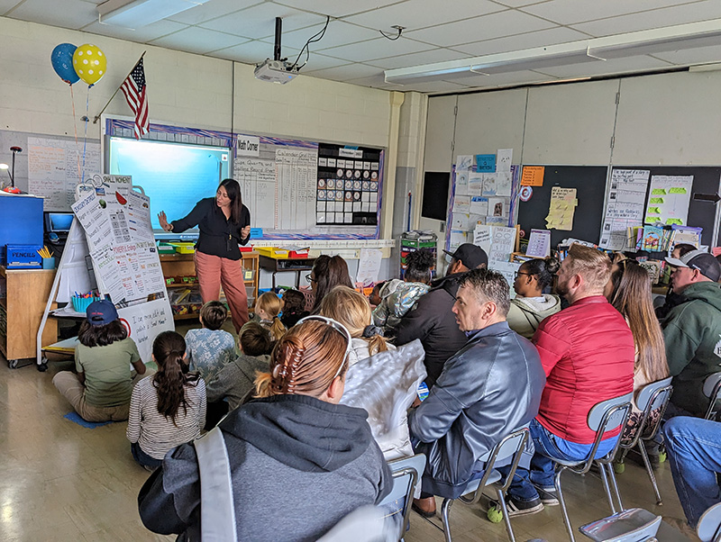 A classroom setting with many adults sitting in chairs, watching a woman at the front of the room talk and write on a large board.