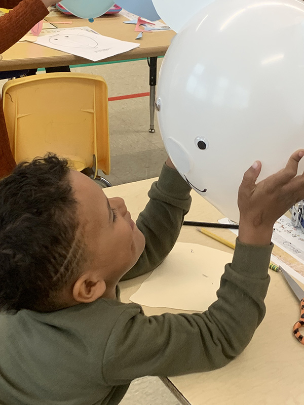 A little boy sits at a desk holding up a white balloon and smiling at it. The balloon has a face drawn on it.