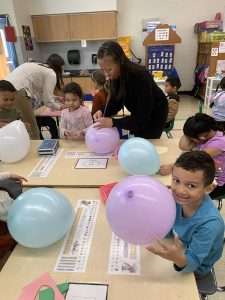 A table of kindergarten kids all with balloons - purple, blue and white. One boy on the end holds up his purple balloon and smiles.