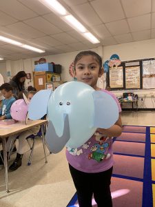 A kindergarten girl wearing a purple shirt holds up a blue balloon that has a trunk and big ears like an elephant.