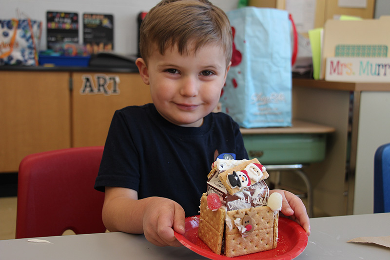 A boy with short light hair, wearing a black shirt, holds a red plate with a gingerbread house made of graham crackers, and decorated with frosting and candy.