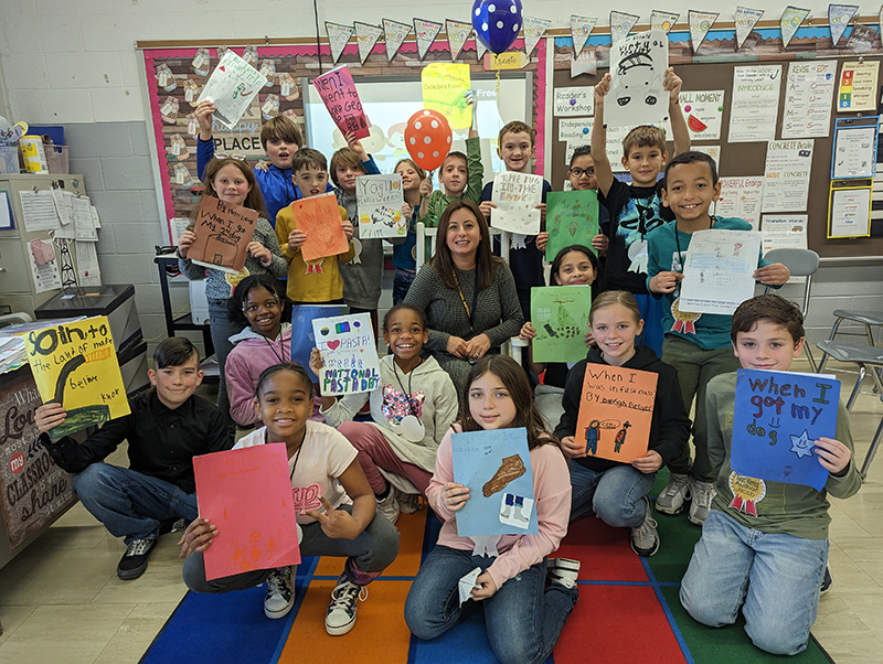 A group of 17 fourth-grade students gathered around their teacher who is sitting. They are all holding the books they've created.