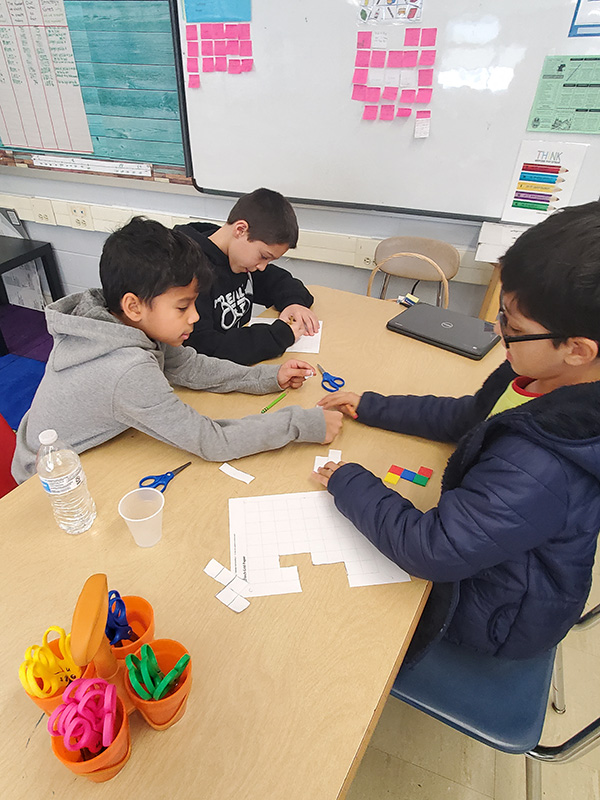 Three fourth-grade boys sit at a table working with small white blocks.