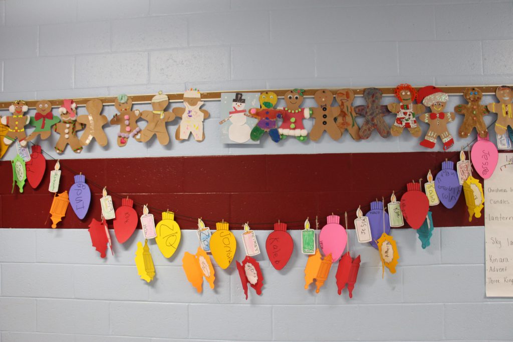 Hanging on a wall is a line of paper gingerbread men. Below them is a clothesline with little clips holding paper cut outs of Christmas bulbs, candles and lanterns.