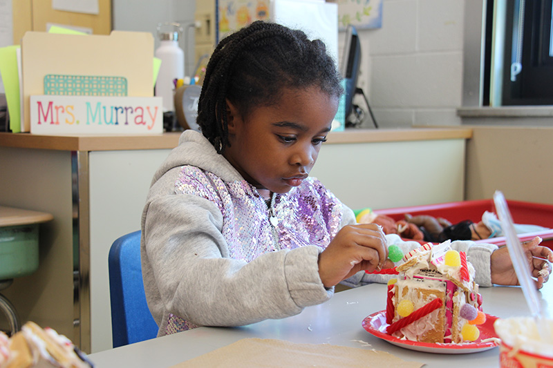 A little girl with dark hair, wearing a gray and purple sweater, decorates a small gingerbread house with candies.