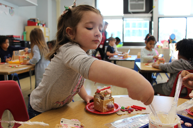 A prek girl with longer brown hair, wearing a gray shirt, reaches with a spreader into a tub of icing for her gingerbread house.