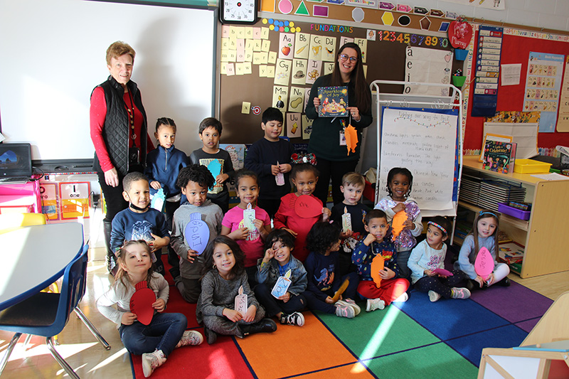 A group of 16 pre-K students sitting, kneeling and standing in three rows on a colorful rug. They are all holding paper candles, lanterns and christmas bulbs and smiling. There are two women in the back, one with long dark hair and glasses holding a book and another with short reddish hair, red shirt and black vest.