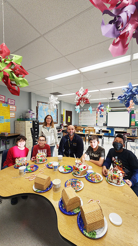 Four sixth-grade students sit at a half circle table with a femals teacher standing behind them and a man sitting with them. Everyone is smiling. There are undecorated gingerbread houses on the table.