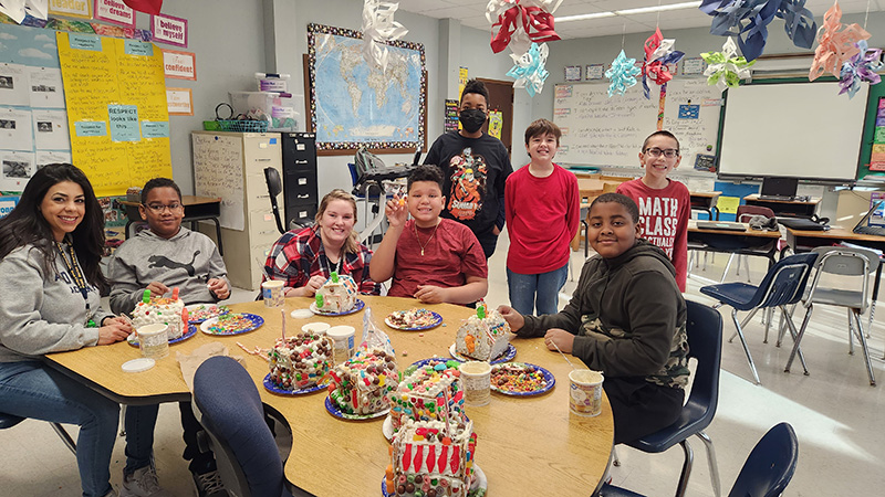 Three sixth-grade boys sit at a half-circle table with two adult women. All are smiling and there are seven gingerbread houses on the table all decorated. Three other sixth-grade boys are standing behind them smiling.