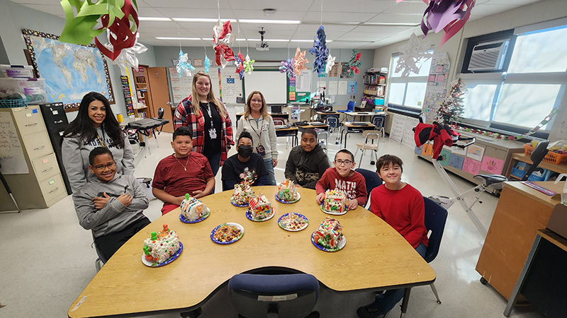 Six sixth-grade boys sit at a semi-circle table with decorated gingerbread houses on the table. There are three female teachers behind them. Everyone is smiling.