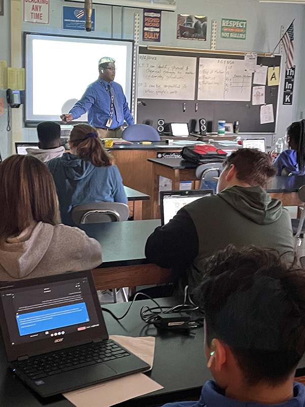 A male teacher stands in front of a classroom of students. He is wearing a blue shirt and is standing in front of a screen. There are students sitting at tables, including one wearing an earbud and watching a screen on a Chromebook.