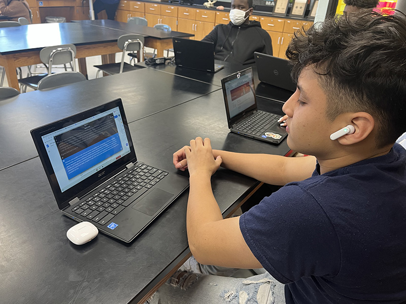 A middle school young man wearing a black tshirt and with short dark hair, sits at a table with his chromebook. He has an earbud in his left ear and is looking at the chromebook.