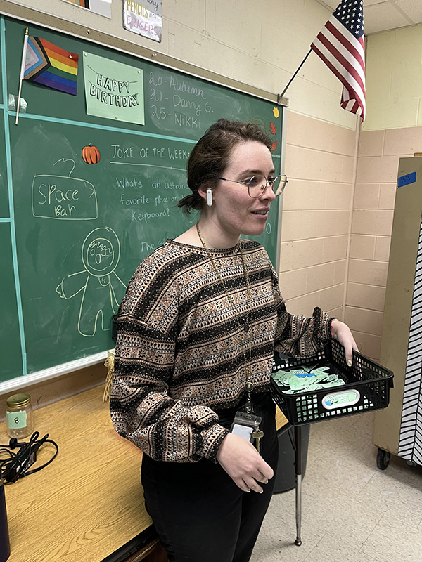 A female teacher stands in front of a green chalkboard. She has an earbud in her ear. She has her dark hair pulled back and is wearing a tan and black sweater.