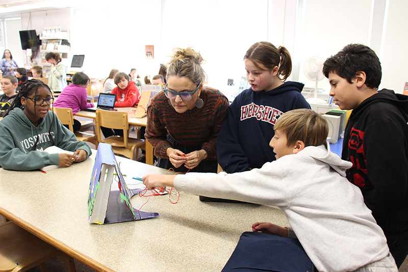 Four students stand around a table with a female teacher. The boy on the right, with sandy hair and a gray sweatshirt on, points to something on the board they are building as the others look at it.
