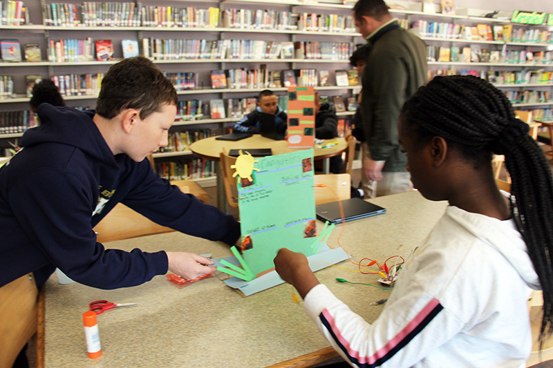 Two students work on a board that has wires attached to it. It is on a table and in the background are shelves with library books on them.