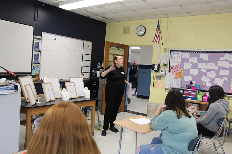 A woman all dressed in black stands in front of a class of eighth-grade students showing them jewelry she has made.