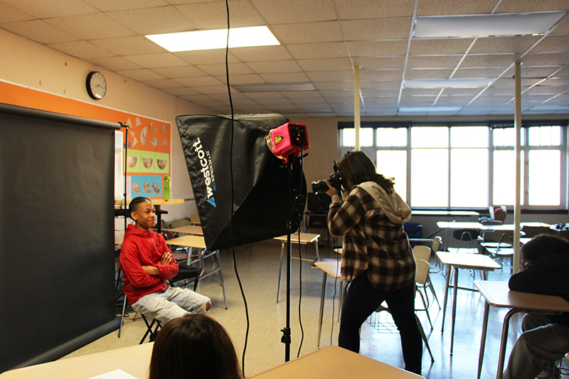 A photographer with long dark hair and wearing a plaid shirt and dark pants, stands behind a camera. There is a young man with a red shirt sitting in front of the camera.