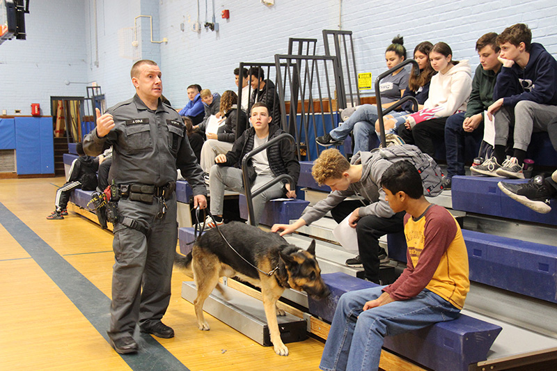 A police officer dressed in a uniform holds the leash to a large German shepherd. The dog is over by many students sitting on bleachers in a gym.