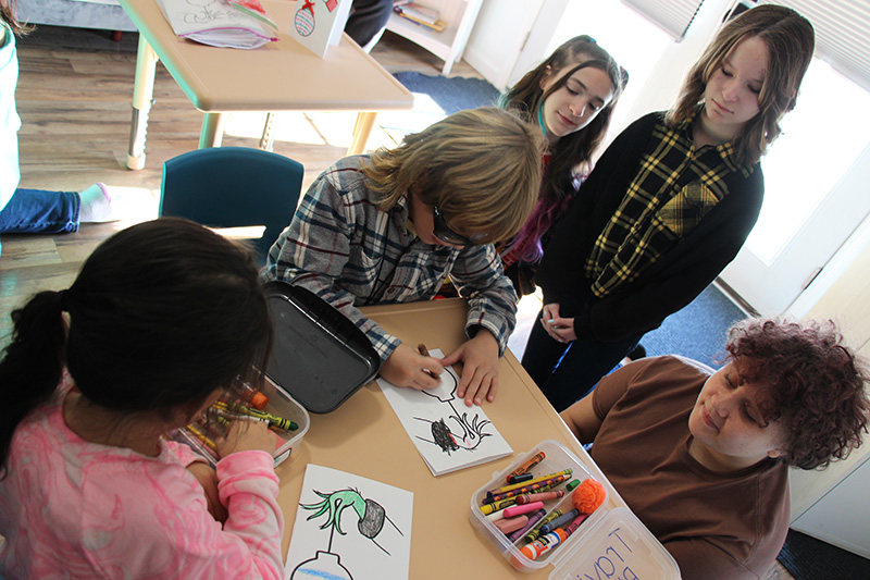 Younger elementary students color pages while older students stand by.