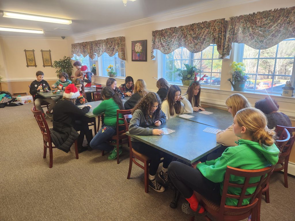 A large group of senior citizens sit at tables playing bingo, with middle school students around them.