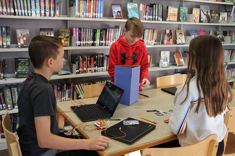 Three middle-school students - two boys and a girl - work together on a project with chromebooks on a table, several different colored wires and a board made of cardboard.