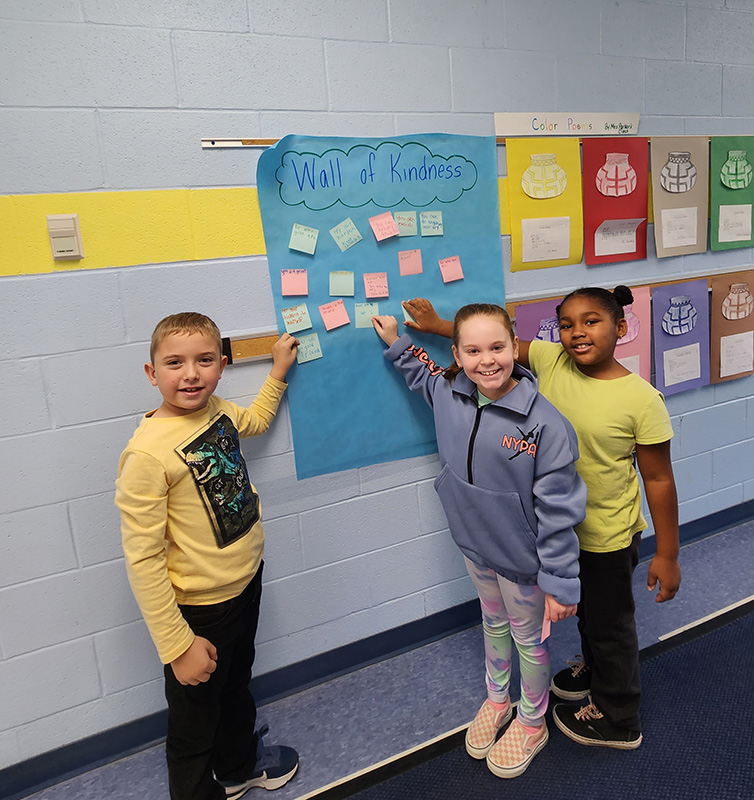 Three younger elementary students stand smiling as they put post-it notes on the wall.