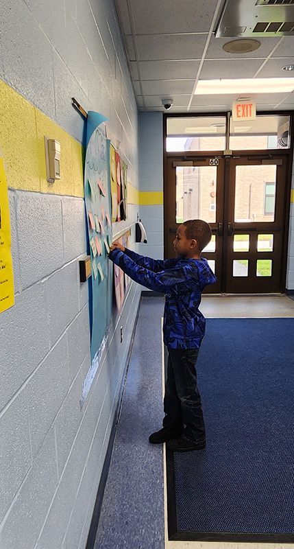 A young elementary student, dressed in blue, puts a message on the positivity wall.
