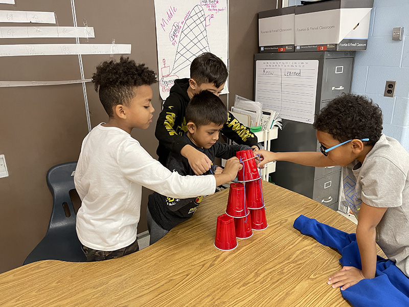Four third-grade boys work together to stack red cups on a desk. They are using string to get the cups in the formation.
