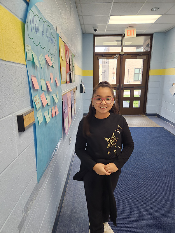 A young girl, wearing glasses and a shirt with stars on it, stands by the positivity board.