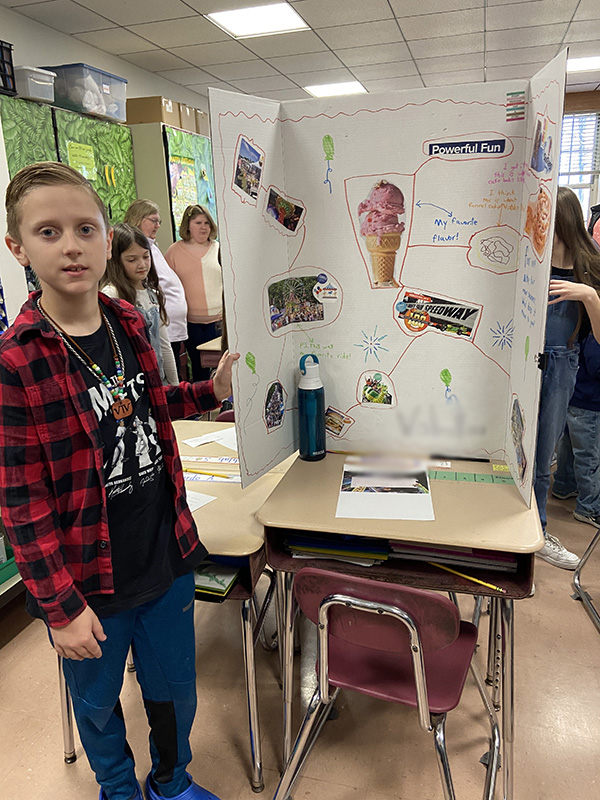 A fourth grade boy with a black and red plaid shirt on stands near his large poster with information about ice cream on it.
