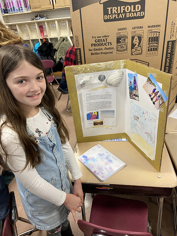 A fourth-grade girl with  long brown hair smiles as she stands next to a desk with her project on it.