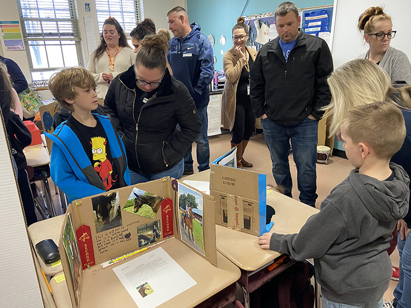 An adult stands with a student as they look at a project on one side of a desk, while another student and adult stand on the other side of the desk looking at another project.