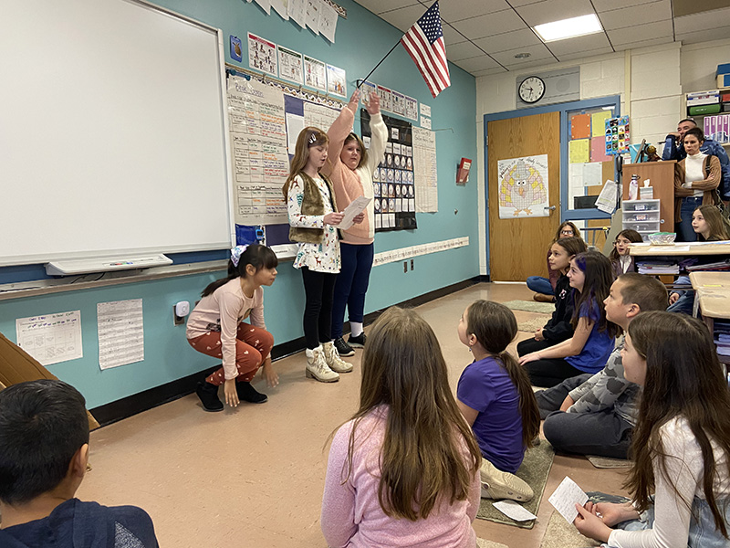 A group of three fourth-grade girls stand in front of the class as other students squat in front of them and adults watch from behind the students.