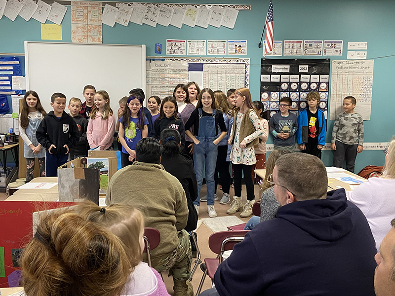 A large group of fourth-grade students in front of a classroom. Adults are sitting in the audience watching them. 