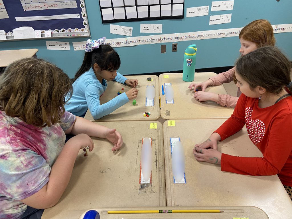 Four elementary students sit at four desks that are pushed together and play dreidel game.