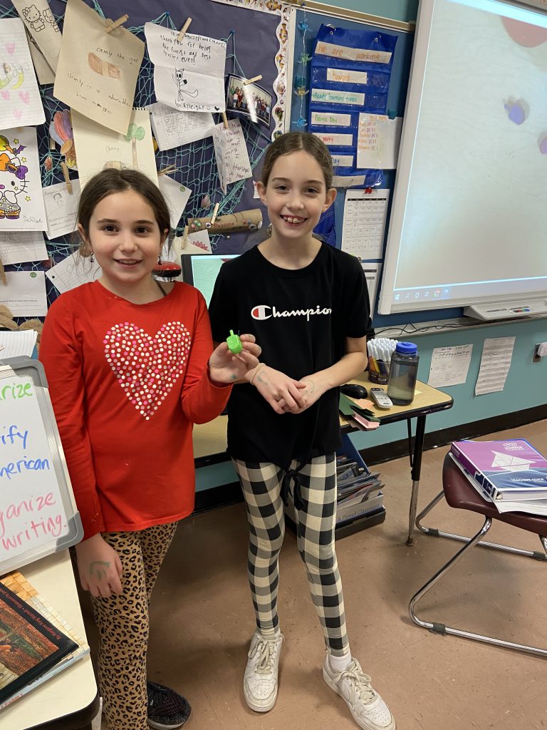 Two fourth grade girls, the girl on the left is wearing a red shirt with a white heart on it and the girl on the right has a black shirt and black and white plaid leggings - stand together holding their dreidel.