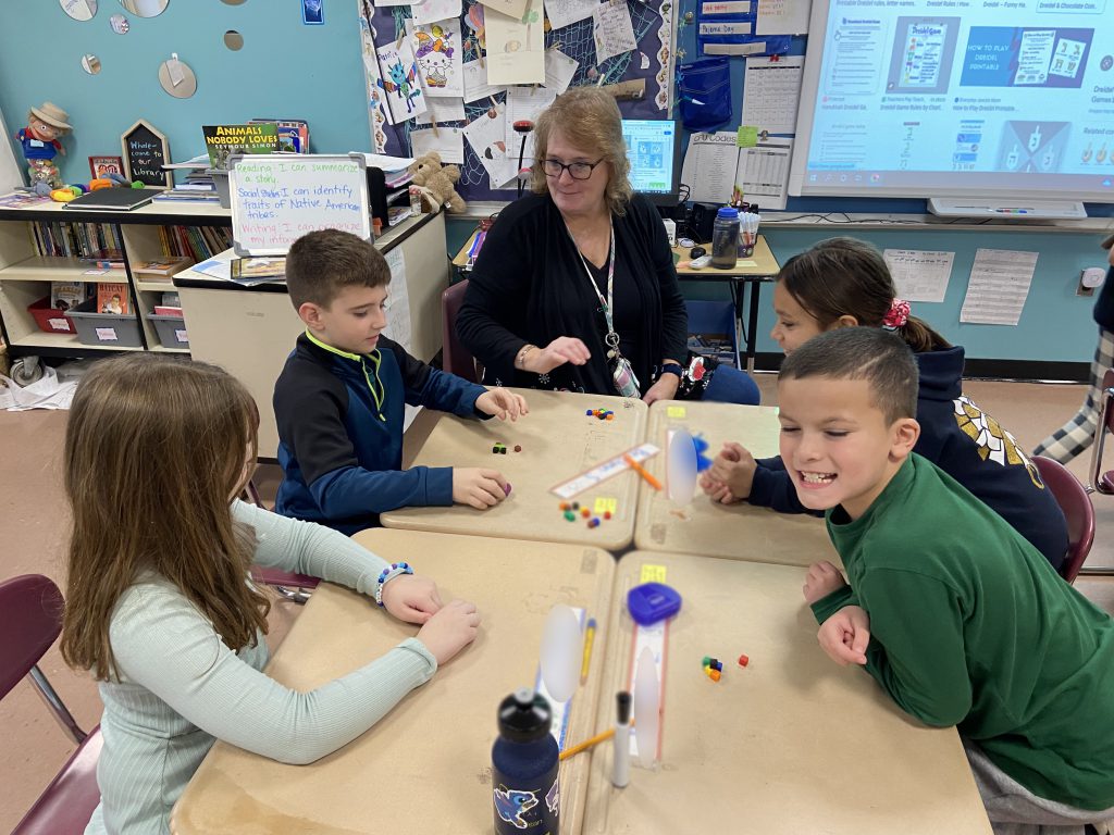 A group of four fourth-grade students smile as they play dreidel with an adult woman.