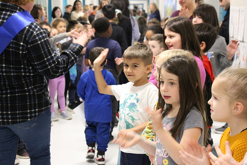 A hallway filled with children and adults as a group of kids walks past them in a parade. A boy holds up his hand for a high-five and a girl with long dark hair claps.