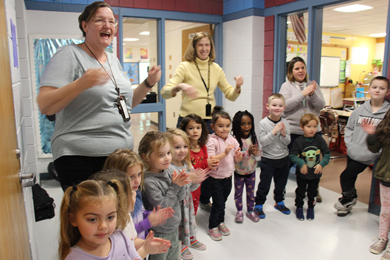 A group of adults and small elementary students stand in a hallway and clap and cheer for kids walking by.