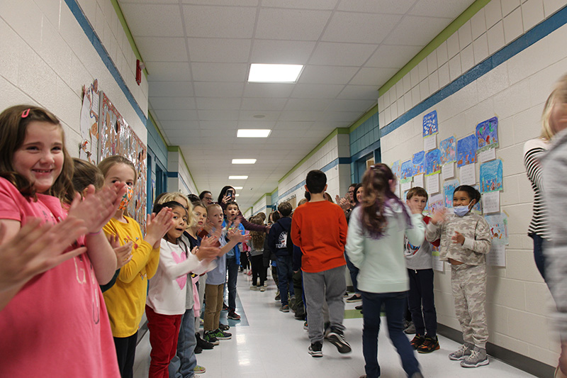 Elementary students line the hallway and clap as other students walk by in a parade.