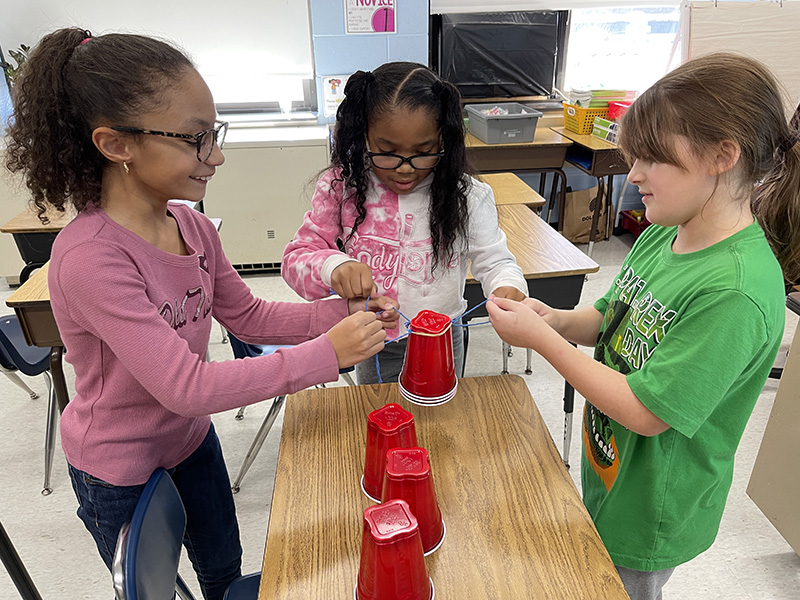 Three third-grade girls work together to stack red cups on a desk. They are using string to get the cups in the formation.