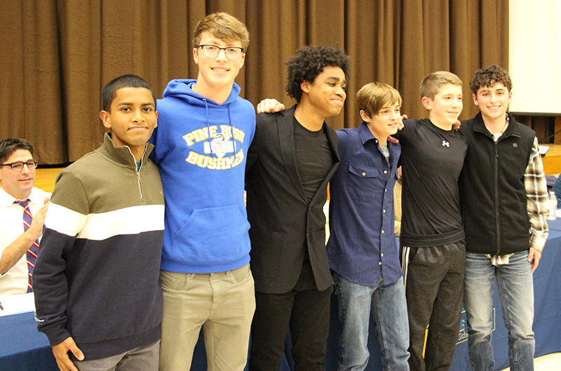 Six high school young men stand arm in arm with a stage and curtain behind them. There is a man sitting to the left clapping.