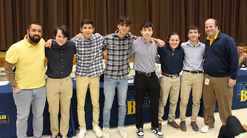 Seven high school age young men stand arm in arm across the front of an auditorium with a man, their coach, at the far right.