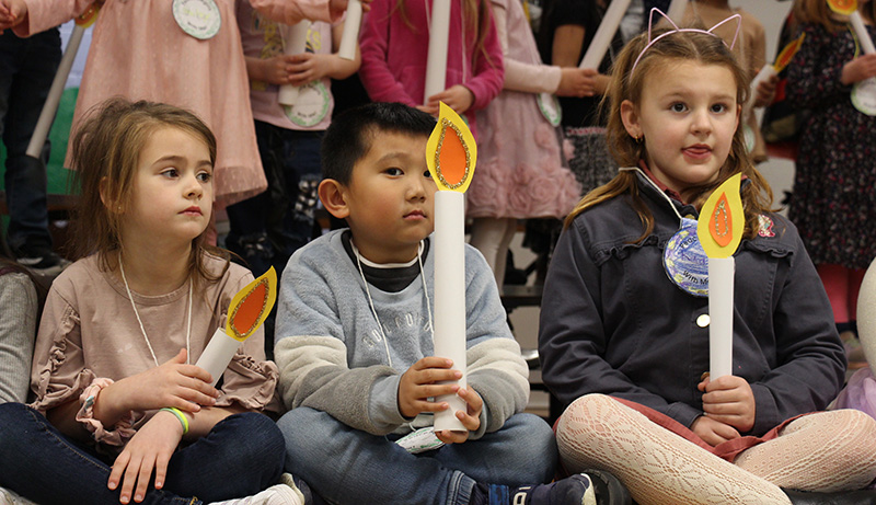 A kindergarten boy, with short dark hair wearing a gray shirt, holds a tall paper candle as he sits on a stage.