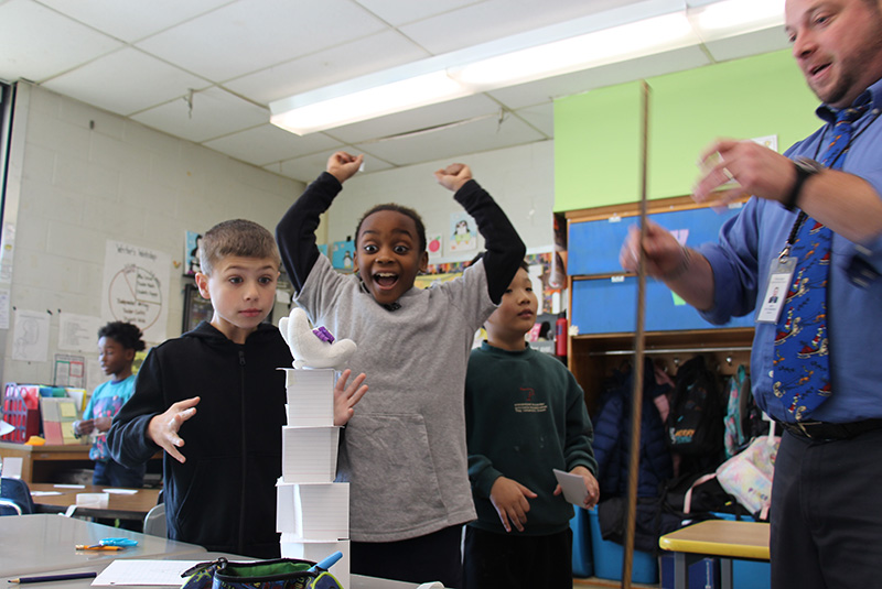 Three third-grade boys stand looking at a structure they built using index cards and tape. One boy's eyes are wide open in amazement, another has his arms up in the air cheering. The third is looking at his teacher who is standing there with a yardstick measuring it.