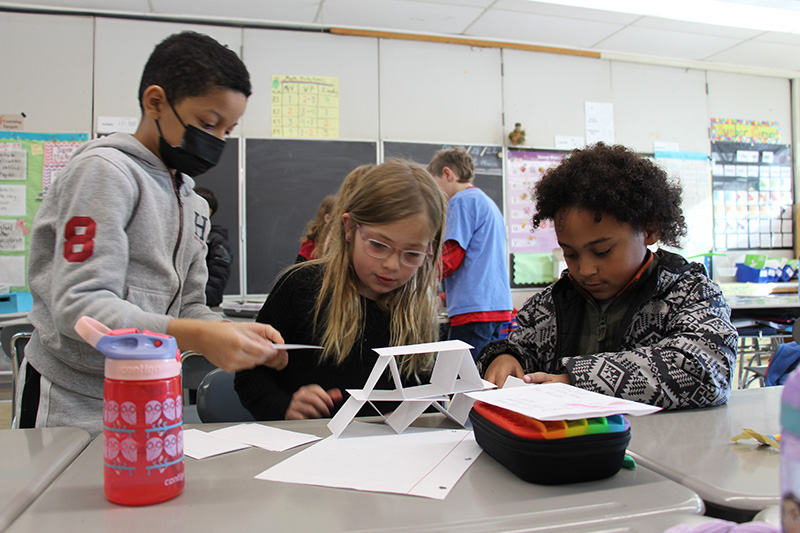 Three third-grade students work at a desk building a structure with index cards.